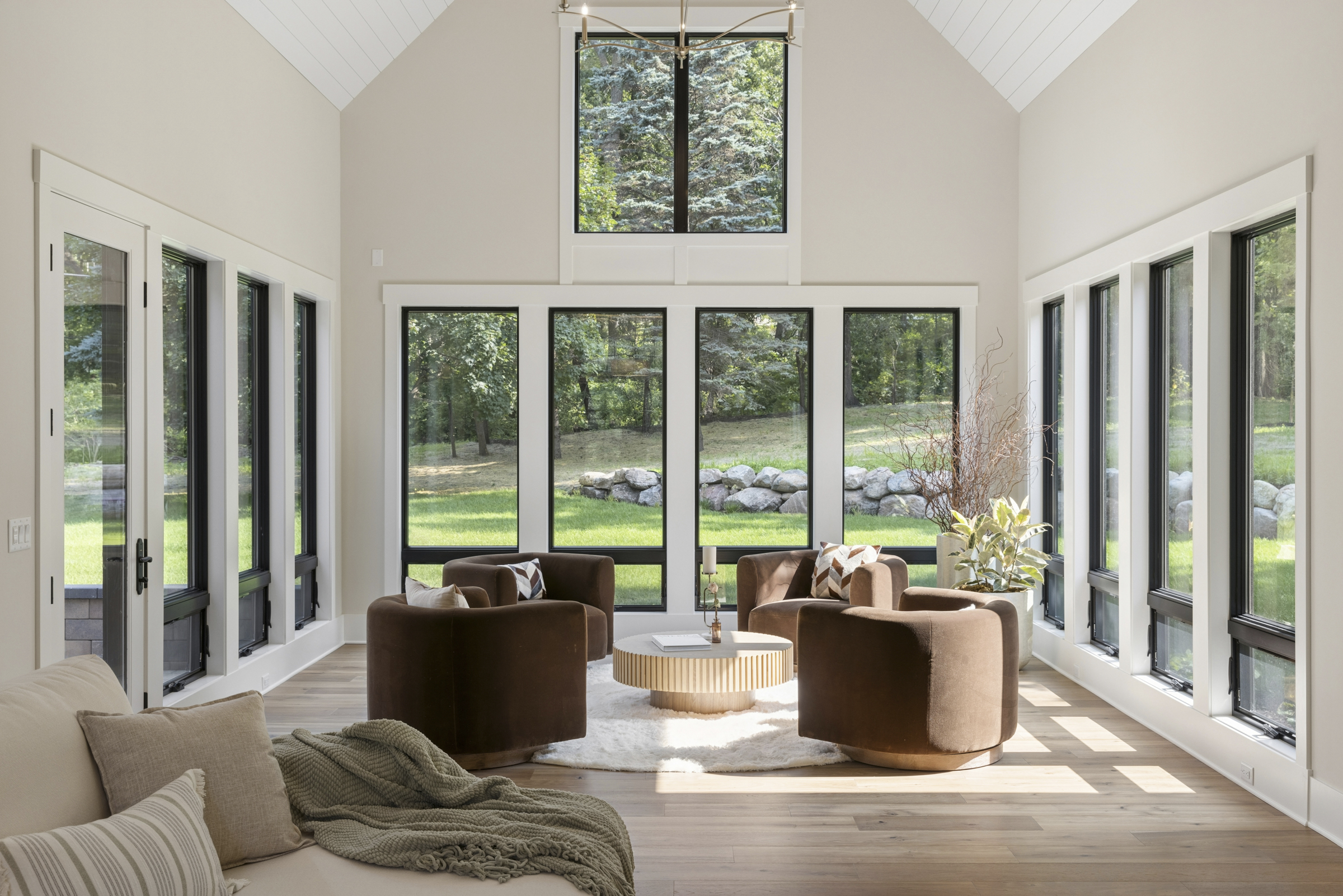 Sunlit great room seating area at 6205 Hillside surrounded by tall black-framed windows overlooking the wooded backyard.