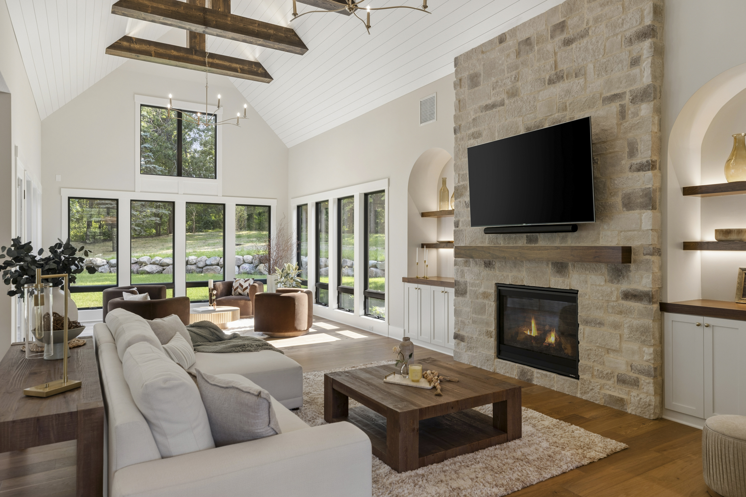Great room at 6205 Hillside with stone fireplace, vaulted ceiling, exposed wood beams, expansive windows, and views of the wooded backyard.