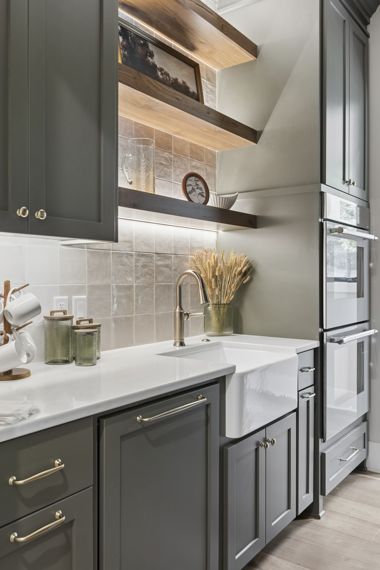Butler's pantry at 6205 Hillside with farmhouse sink, dark custom cabinetry, brass hardware, tile backsplash, illuminated wood shelves, and built-in ovens.