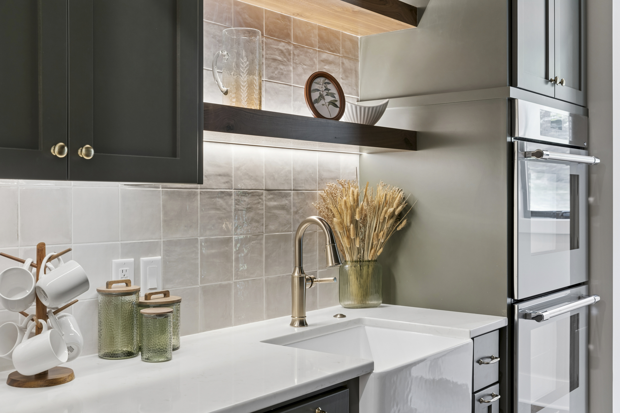 Detailed view of the 6205 Hillside butler's pantry with farmhouse sink, illuminated floating shelves, tile backsplash, custom cabinetry, and built-in ovens.