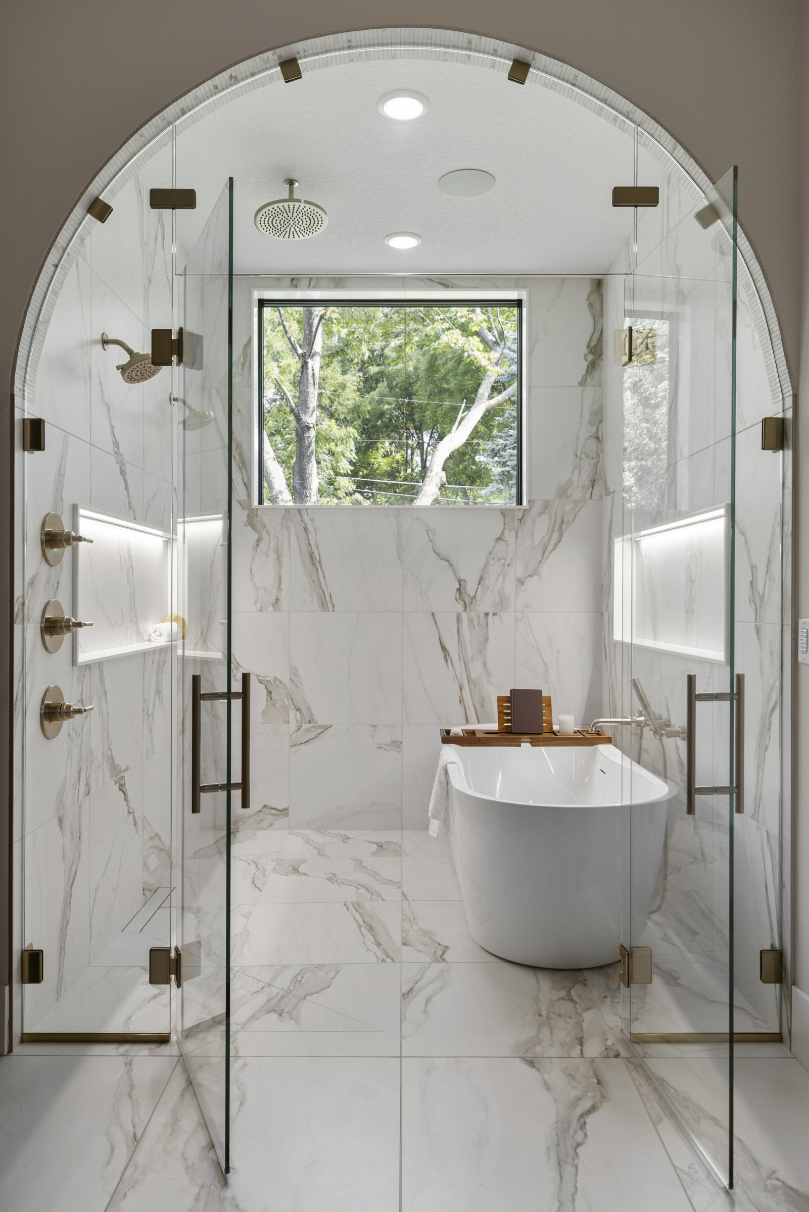 Luxury primary bathroom at 6205 Hillside with arched wet room, glass shower enclosures, freestanding soaking tub, marble-look tile, and brass fixtures.