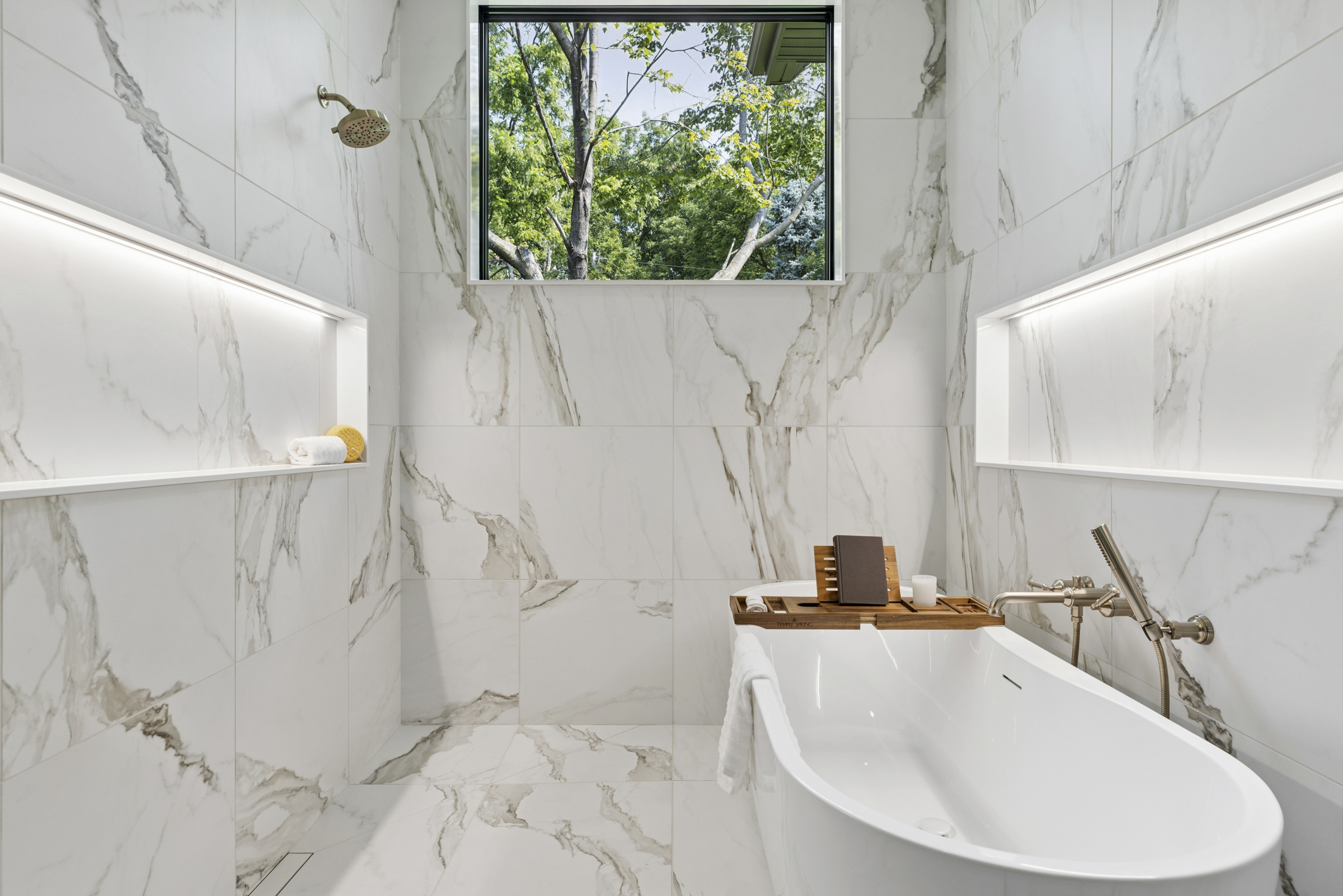 Primary bathroom wet room at 6205 Hillside with freestanding soaking tub, marble-look tile, illuminated shower niches, and large window overlooking mature trees.