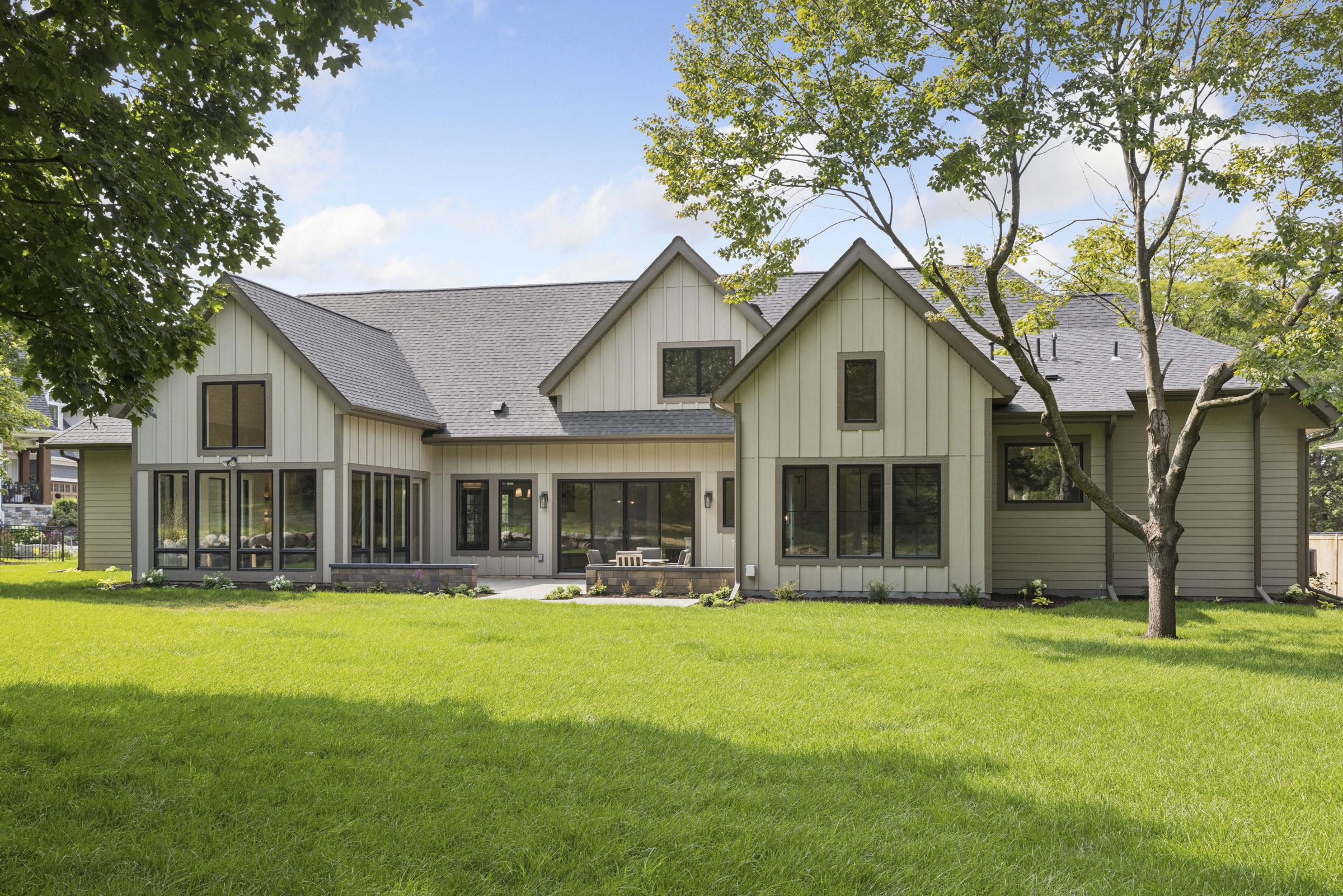 Wide rear exterior view of the 6205 Hillside custom home with multiple gables, black-framed windows, patio, landscaping, and spacious backyard.