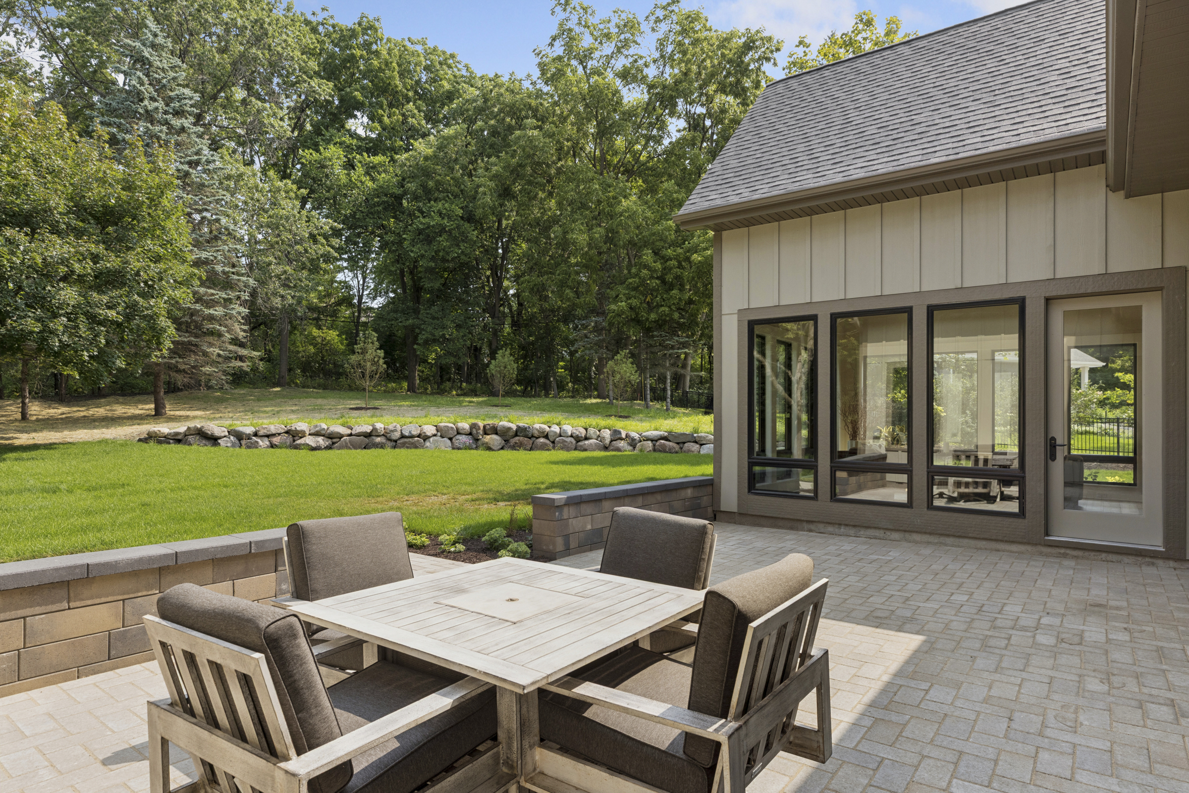 Backyard patio at 6205 Hillside with outdoor dining area, paver hardscape, landscaped retaining wall, expansive lawn, and mature wooded surroundings.